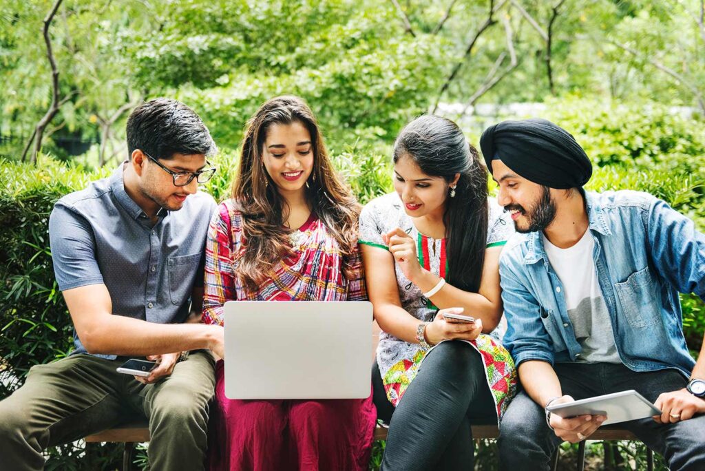 Group of young students sitting outdoors, collaborating on a laptop and tablet during a study session.
